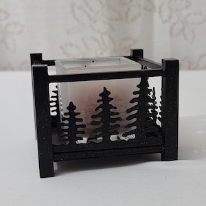 May include: A black metal candle holder with a rectangular frame. The sides feature a cut-out forest design. A glass insert holds a lit candle, casting a warm glow. The candle holder sits on a white surface.