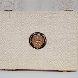 May include: A rectangular, off-white game case with a textured surface. The case has a gold-colored metal emblem in the center with the words "MAGNETIC BOOKSHELF BACKGAMMON" and the number "1863". The case is closed with gold-colored hinges.