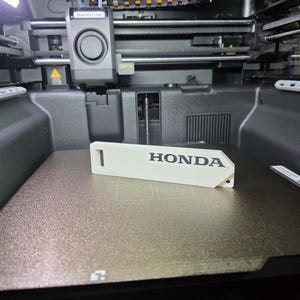 May include: A white 3D printed rectangular keychain tag featuring the word HONDA embossed in black lettering. The tag has a small rectangular cutout on the left side and a circular hole on the right side, resting on a textured 3D printer bed.