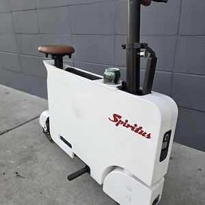 May include: White Honda Spiritus scooter with a brown seat and handlebars. The scooter has black accents and a red "Spiritus" logo. A can of beverage sits in a small compartment.