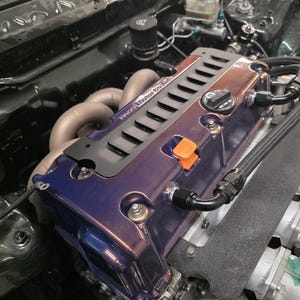May include: A close-up of a car engine bay. The engine features a purple and bronze valve cover with a black accent. The engine bay is dark green. The image shows various engine components, including pipes and a black air filter.