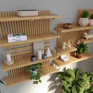 Puede incluir: Estanterías de pared de madera clara, dispuestas en un diseño moderno. Las estanterías exhiben objetos decorativos como plantas, libros, velas y pequeñas esculturas. El diseño incorpora listones de madera verticales.
