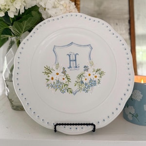 Könnte beinhalten: Ein weißer Keramikteller mit gewelltem Rand und blau gepunkteter Verzierung. In der Mitte befindet sich ein blaues Wappen mit dem Buchstaben H, flankiert von handgemalten Blumenarrangements in Weiß, Gelb, Grün und Hellblau.