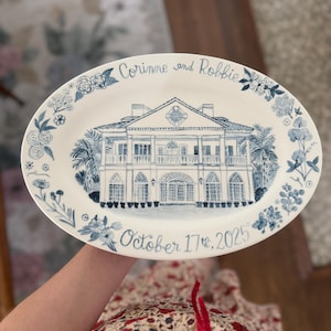 Puede incluir: Plato ovalado de cerámica con una ilustración pintada a mano de una casa de dos pisos en azul y blanco. El plato está decorado con detalles florales y los nombres "Corinne y Robbie" y la fecha "17 de octubre de 2025".