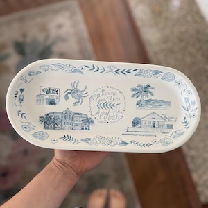 May include: An oval, white ceramic serving platter with blue illustrations. The platter features drawings of buildings, a crab, palm trees, and the text "Catherine and Ben April 4th 2026". Decorative floral and seashell elements surround the illustrations.