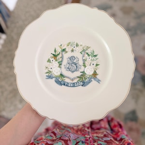 May include: A white decorative plate with a scalloped edge and gold trim. The plate features a floral wreath design with a blue ribbon and the date "9-26-2025". A blue letter "S" is in the center of the wreath.