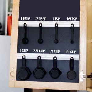 May include: A black chalkboard with white labels for measuring spoons and cups. The labels read: 1 TBSP, 1/2 TBSP, 1 TSP, 1/4 TSP, 1 CUP, 3/4 CUP, 1/2 CUP, 1/4 CUP. Black measuring spoons and cups are hanging on a white metal rack.