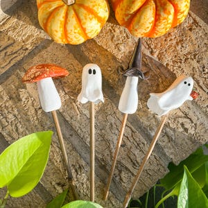 May include: Two orange and yellow pumpkins sit above four Halloween-themed garden stakes. The stakes feature a mushroom, a ghost, a witch hat, and a ghost holding a small orange. The stakes are on wooden dowels.