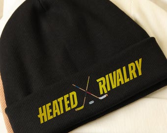 May include: Black knit beanie with the words "HEATED RIVALRY" in yellow, along with crossed hockey sticks. The beanie is folded at the brim and rests on top of other beanies in cream and tan.