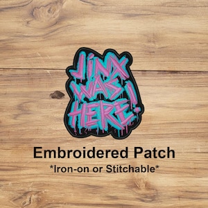 Peut inclure: Écusson brodé avec le texte "Jinx Was Here!" dans un style graffiti. Le texte est rose avec un contour turquoise, sur un fond noir. L'écusson est posé sur une surface en bois.