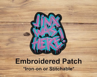 May include: Embroidered patch with the text "Jinx Was Here!" in a graffiti style. The text is in pink with a turquoise outline, set against a black background. The patch is on a wooden surface.