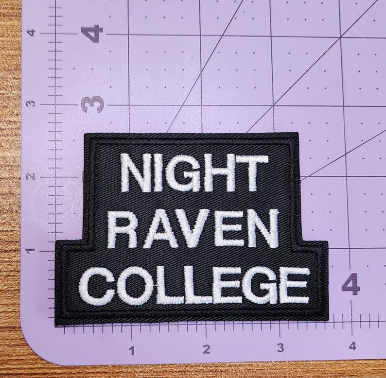 May include: Black rectangular patch with white embroidered text reading "NIGHT RAVEN COLLEGE". The patch is approximately 10 cm wide and 6.3 cm tall. The patch is on a purple cutting mat.
