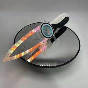 May include: A futuristic-looking dagger with a white and black handle, a silver blade, and a circular guard with blue accents. The blade has a holographic, iridescent effect, reflecting orange and yellow light. The dagger rests on a reflective surface.