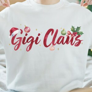 Op de afbeelding: Een wit sweatshirt met de rode tekst Gigi Claus in een sierlijk lettertype. Het ontwerp bevat een rode kerstmuts, roze en groene kerstballen, een kleine rode strik, groene hulstblaadjes met rode bessen en kleine gouden sterretjes.