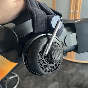 Könnte beinhalten: Schwarzes VR-Headset mit einem dunkelgrauen Metallarm und einem schwarzen Mesh-Lautsprecher. Das Headset hat ein gepolstertes Kopfband und Ohrmuscheln. Der Markenname "Pimax" ist auf dem Lautsprecher sichtbar.