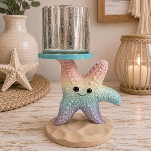 May include: A candle holder featuring a pastel rainbow-colored starfish base with a smiling face, standing on a textured sand-colored circular platform. A teal disc sits atop the starfish, supporting a cylindrical, vertical-ribbed mercury glass cup.