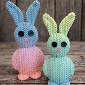 May include: Two knitted bunny figurines. One is blue and pink, the other is green and yellow. Both have button eyes and pink noses. The bunnies are standing on a wooden surface.