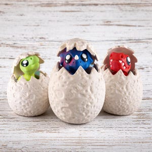 May include: Three ceramic dinosaur egg figurines, each with a different coloured dinosaur emerging. The dinosaurs are green, blue, and red. The eggs are off-white with a textured surface, and the background is light wood.