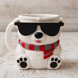 May include: A white ceramic mug shaped like a polar bear wearing black sunglasses and a red and gray scarf. The mug has a handle and paw-shaped feet. The bear's nose is black, and its mouth is pink.