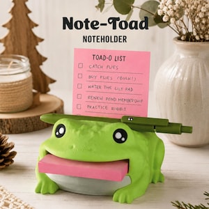 May include: A bright green frog-shaped note holder with black and white eyes. It holds a pink sticky note with the text TOAD-O LIST, CATCH FLIES, BUY FLIES (BULK!), WATER THE LILY PAD, RENEW POND MEMBERSHIP, PRACTICE RIBBIT. A green pen rests on top.