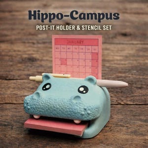 Peut inclure: Un porte-bloc-notes en forme d'hippopotame bleu avec un bloc-notes rose et un stylo. Un calendrier rose pour janvier est en arrière-plan. Le texte "Hippo-Campus POST-IT HOLDER & STENCIL SET" est en haut.