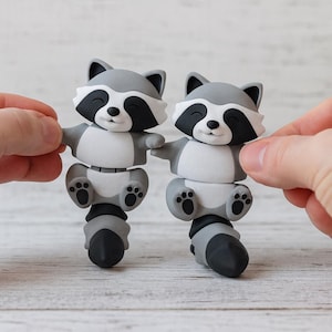 May include: Two gray and white raccoon figurines with black accents. The raccoons have their arms outstretched and are smiling. The figurines have a segmented body and a black and gray striped tail. The background is a light wood surface.