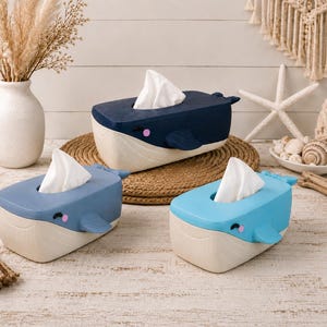 May include: Three whale-shaped tissue box covers in dark blue, medium blue, and light blue. Each container features a cream-colored base, a top opening for tissues, side fins, and a small tail. The whale faces have closed eyes and pink cheeks.