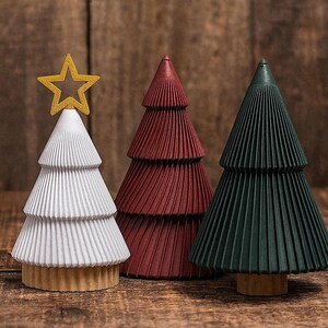 May include: Three decorative Christmas trees in white, burgundy, and dark green. The white tree has a gold star on top. The trees have a pleated design and are set on a wooden surface against a wooden background.