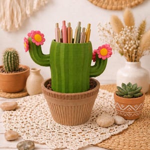 May include: A green cactus-shaped pencil holder with two arms, each featuring a pink flower with a yellow center. The cactus sits in a tan, ribbed pot and contains several metallic pens. The design mimics a potted plant with vertical ridges.