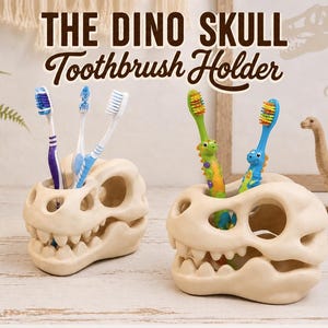 May include: Two off-white dinosaur skull toothbrush holders with hollow eye and nasal cavities for storage. The text THE DINO SKULL Toothbrush Holder is displayed above. Several colorful toothbrushes are inserted into the top openings.