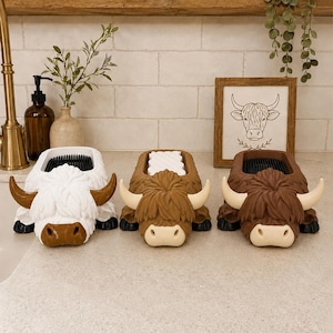 May include: Three highland cow-shaped sponge holders in white, tan, and dark brown. Each holder features a textured, shaggy-look exterior, curved horns, and a rectangular opening on top containing a sponge for kitchen or bathroom utility.