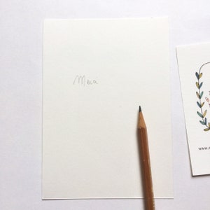 May include: A blank white card with the word "Merci" written in pencil. A wooden pencil with a black eraser is lying on the card.