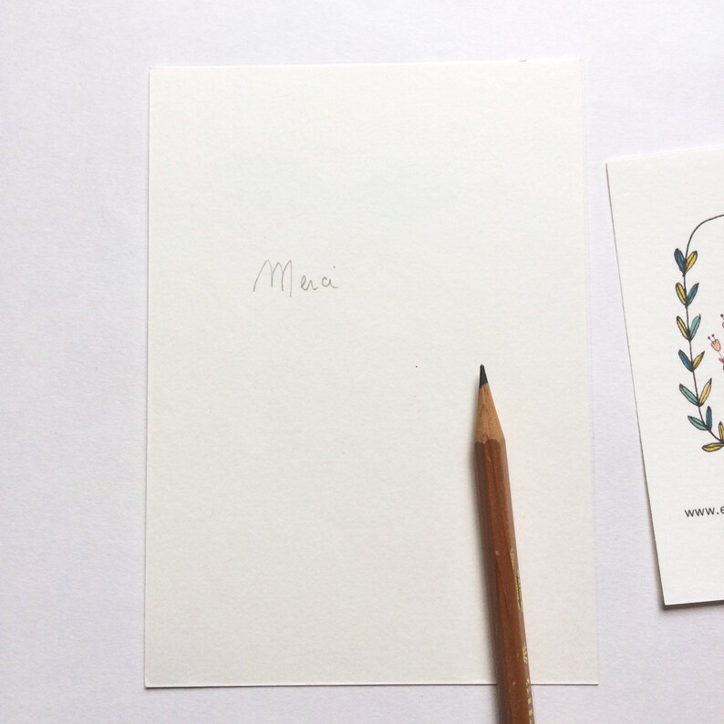 May include: A blank white card with the word "Merci" written in pencil. A wooden pencil with a black eraser is lying on the card.