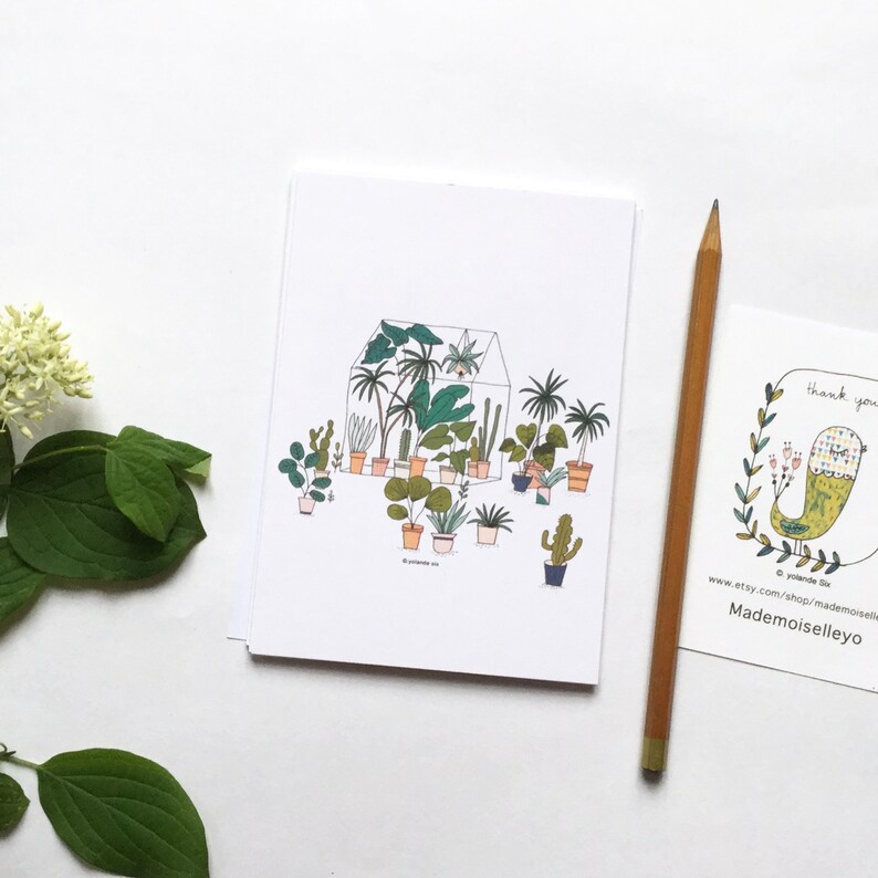 May include: A white card with a hand-drawn illustration of a greenhouse filled with potted plants. The plants are drawn in various shades of green, brown, and orange. The greenhouse is outlined in black and has a white roof. The card has the text "Mademoiselleyo" at the bottom.