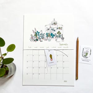 Könnte beinhalten: Ein weißer Kalender für 2026 mit einer botanischen Illustration eines mit Pflanzen gefüllten Gewächshauses. Der Kalender zeigt den Monat September und ein kleines Etikett mit einer Kaktusillustration. Ein Bleistift und eine Dankeskarte sind ebenfalls sichtbar.