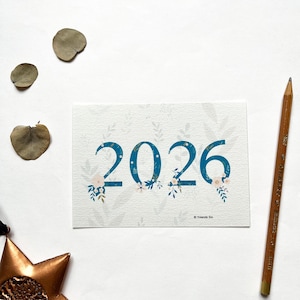 Könnte beinhalten: Eine Grußkarte mit der Jahreszahl 2026 in Türkisblau, verziert mit floralen Akzenten. Die Karte liegt auf einem weißen Hintergrund, zusammen mit einem Bleistift, getrockneten Blättern und einem sternförmigen Ornament. Der Name des Künstlers ist ebenfalls zu sehen.