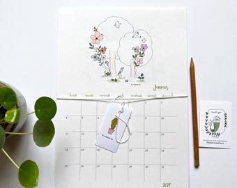 May include: A 2027 wall calendar for the month of Janvier featuring a line art illustration of trees, flowers, and birds. A small white tag with a cactus illustration hangs from a string. A wooden pencil and a small thank you card are included.