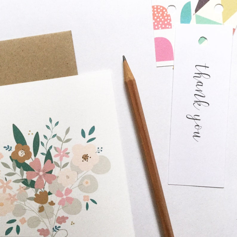 May include: A white card with a floral illustration in shades of pink, brown, and green. The card is blank and has a brown envelope next to it. A pencil and a white card with the text "thank you" are also in the image.