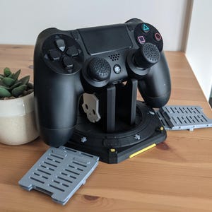 May include: A black PlayStation 4 controller is mounted on a black charging stand with a yellow accent. The controller has two grey thumbsticks with black caps. The stand has two grey, rectangular charging pads on either side.
