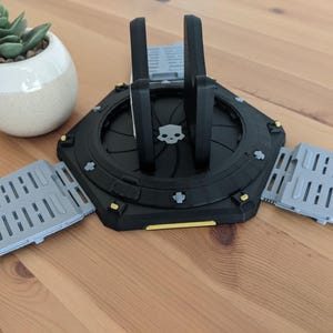 May include: A black and grey 3D printed phone stand with a skull design. The stand has three foldable legs that can be extended to create a stable base.