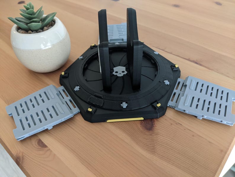 May include: A black and grey 3D printed phone stand with a skull design. The stand has three foldable legs that can be extended to create a stable base.