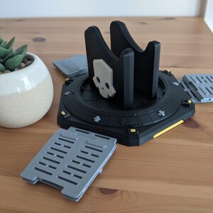 May include: A black 3D printed stand with a skull design and yellow accents. The stand has four grey, rectangular platforms attached to it.