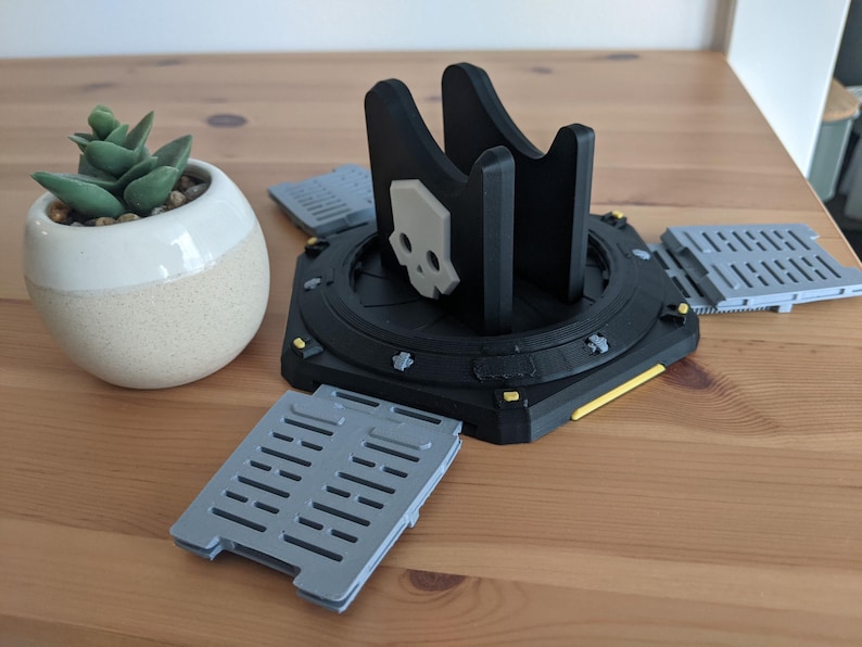May include: A black 3D printed stand with a skull design and yellow accents. The stand has four grey, rectangular platforms attached to it.