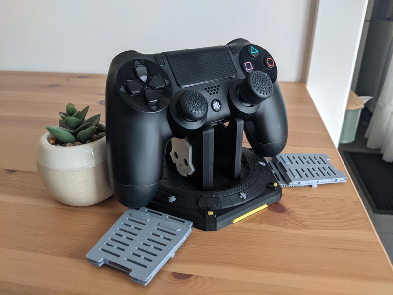 May include: A black PlayStation 4 controller is mounted on a black charging stand with a yellow accent. The controller has two grey thumbsticks with black caps. The stand has two grey, rectangular charging pads on either side.