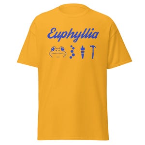 Puede incluir: Una camiseta amarilla con el texto "Euphyllia" en letras azules cursivas. Debajo del texto hay una rana azul, tres puntos azules, una antorcha azul y un martillo azul.