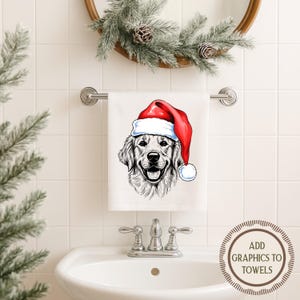 Op de afbeelding: Witte handdoek met een gedetailleerde illustratie van een Golden Retriever met een rode kerstmuts. De handdoek hangt aan een zilveren stang boven een witte wastafel. Boven de handdoek hangt een decoratieve krans.
