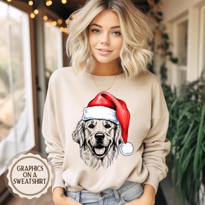 Op de afbeelding: Beige sweatshirt met een afbeelding van een Golden Retriever met een kerstmuts. De hond is afgebeeld in zwart-wit, met een rood-witte kerstmuts. De tekst "GRAPHICS ON A SWEATSHIRT" is gedrukt op een cirkelvormig label.