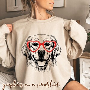 May include: A beige sweatshirt with a graphic of a Golden Retriever wearing red heart-shaped glasses. The dog is smiling. The text "graphics on a sweatshirt" is at the bottom of the image.