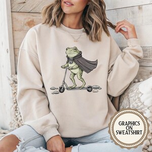 K&ouml;nnte beinhalten: Beiges Sweatshirt mit einem Cartoon-Frosch, der einen Umhang tr&auml;gt und einen Roller f&auml;hrt. Der Frosch ist gr&uuml;n mit einem schwarzen Umhang und einem silbernen Emblem. Das Sweatshirt hat den Text "Graphics on Sweatshirt".