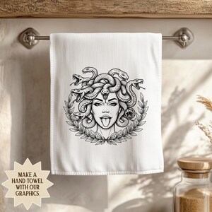 May include: White hand towel featuring a black and white illustration of Medusa with snakes for hair, surrounded by a laurel wreath. The towel is hanging on a metal bar. Text on the bottom left says "MAKE A HAND TOWEL WITH OUR GRAPHICS."